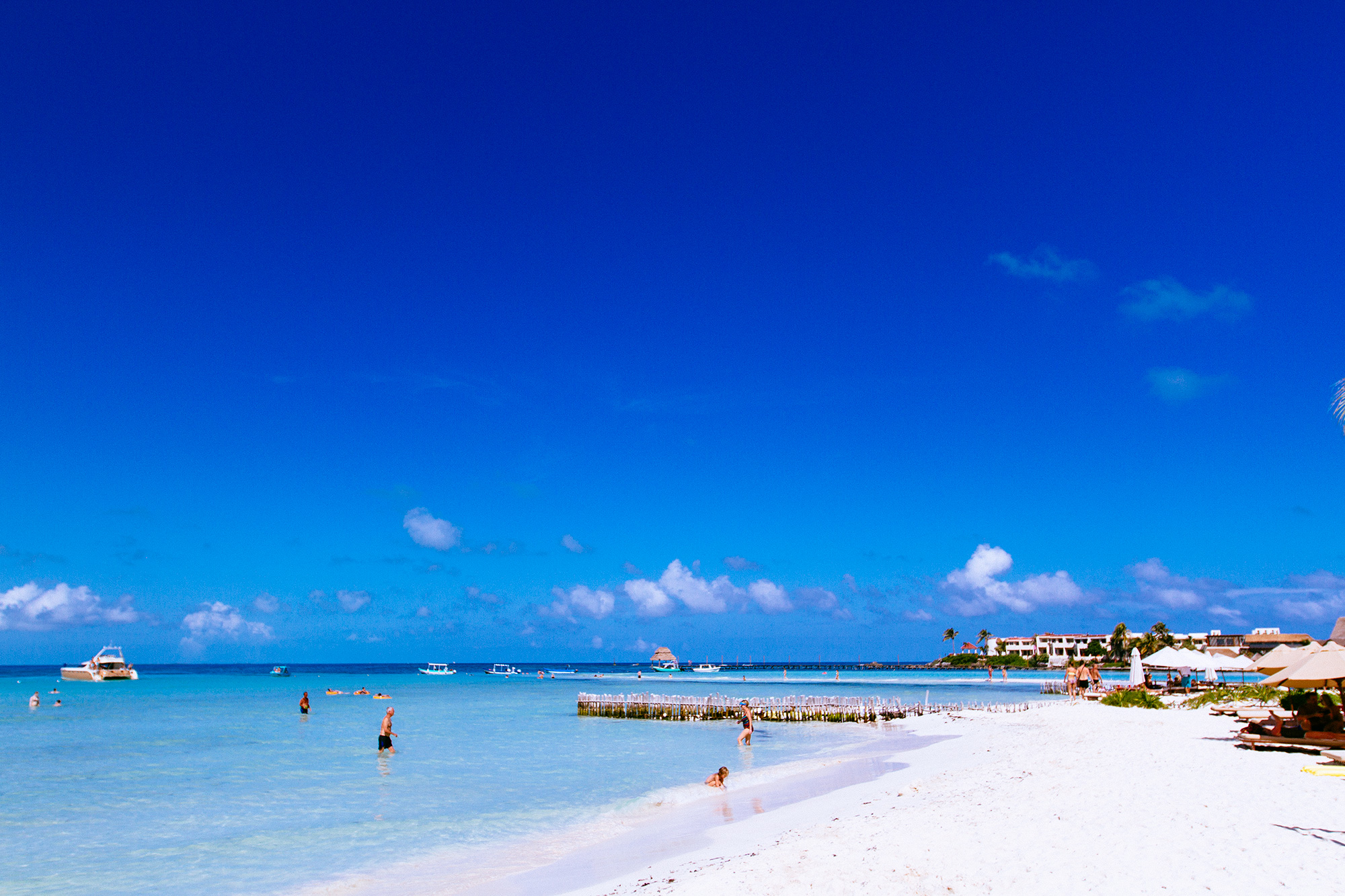 Wide tropical beach with turquoise water, white sand, distant buildings, and boats under a bright sky