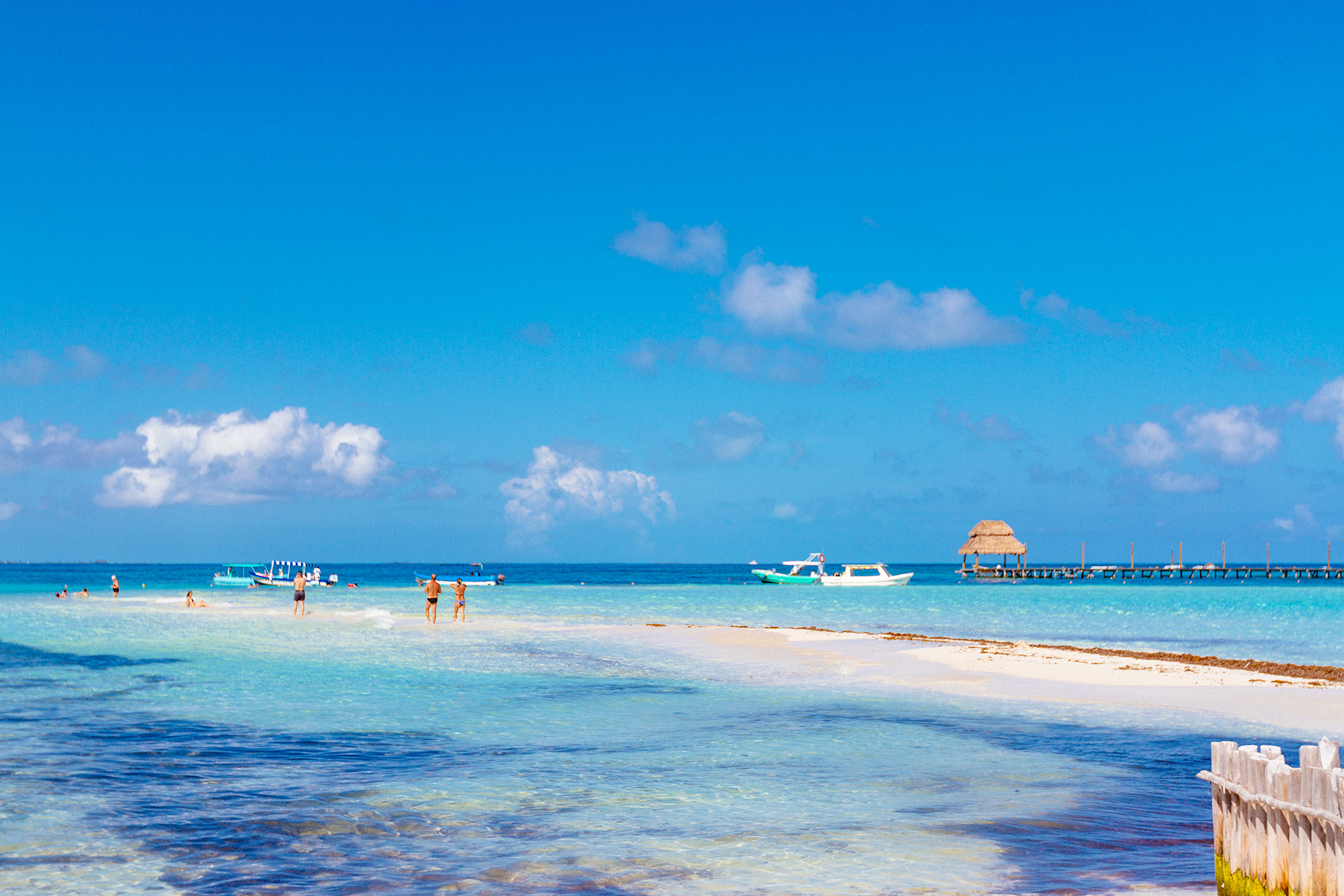 Tropical seascape with turquoise water, a sandbar, small boats, and a thatched-roof structure over the water, bright and serene
